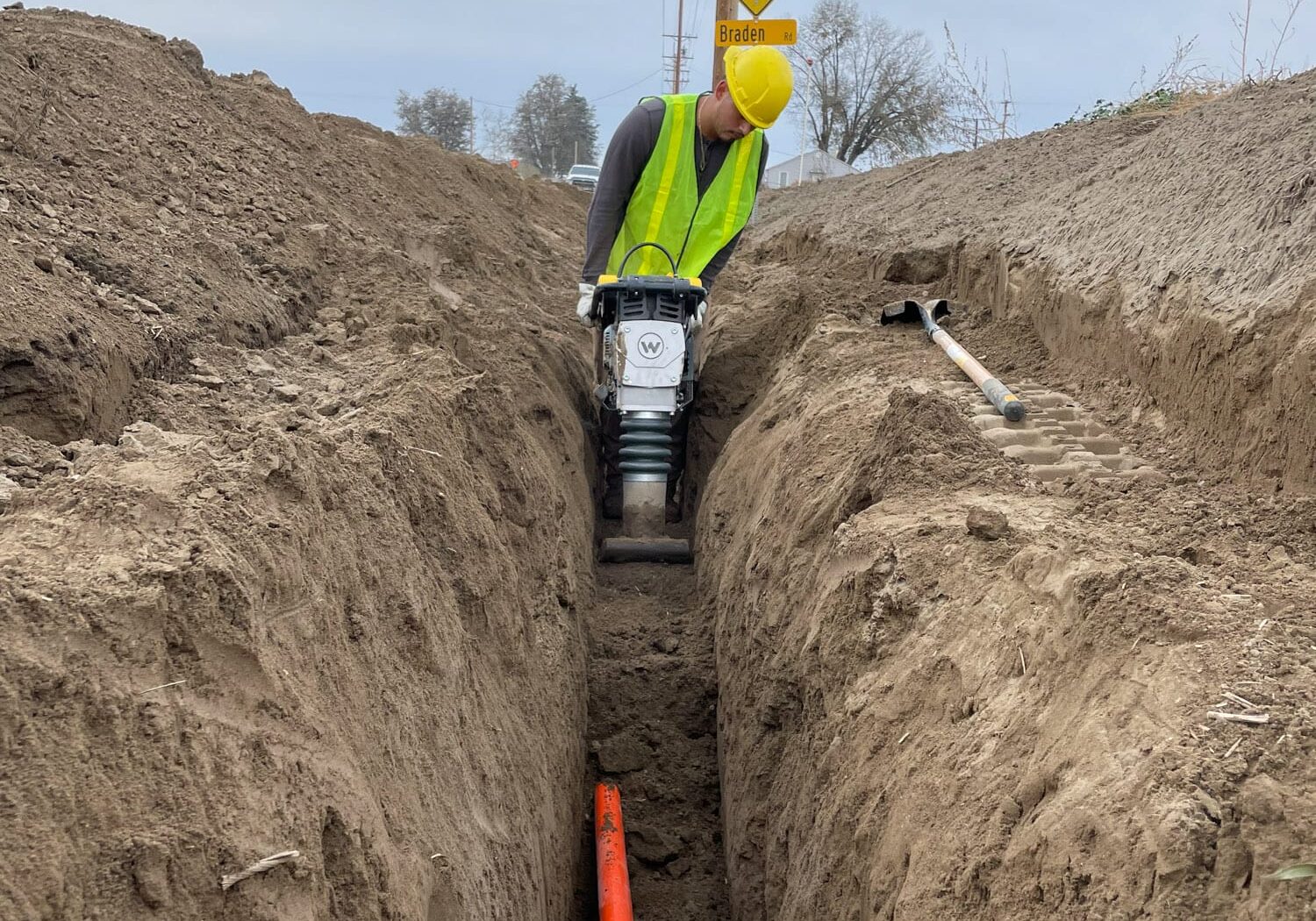 Construction worker compressing soil in trench with orange pipe.